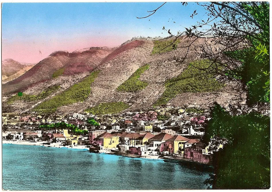 Hand-tinted postcard of a coastal village, pink mountainside rising behind yellow houses